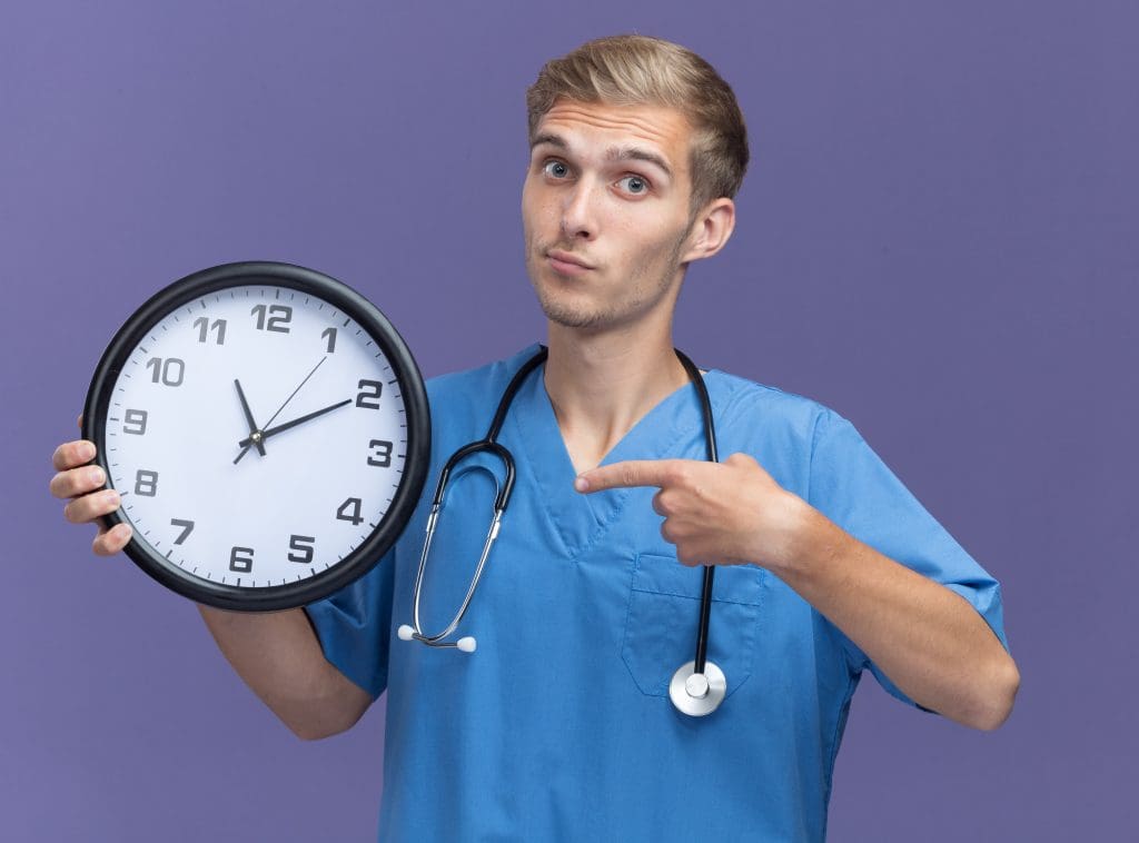 impressed young male doctor wearing doctor uniform with stethoscope holding points wall clock isolated blue wall LIV Hospital