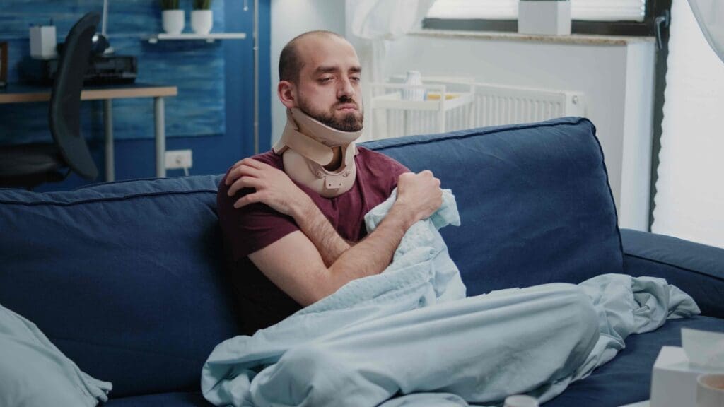 A middle-aged person lying down on a couch with a pained expression, their neck wrapped in a bandage or medical brace