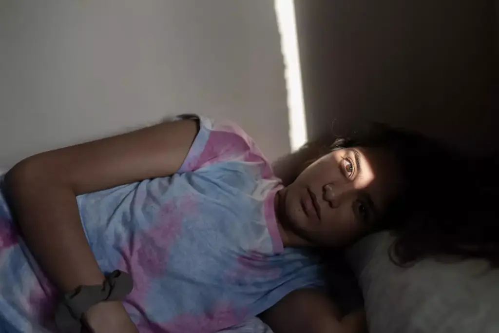 A person lies in bed, their face gaunt and pale from the effects of chemotherapy. Thin wisps of hair cling to their head, a stark contrast to the vibrant life they once held. The room is dimly lit, casting long shadows across the bedsheets, creating a somber and introspective atmosphere. The subject's gaze is distant, conveying the exhaustion and uncertainty of their journey. The scene is captured with a soft, muted color palette, emphasizing the delicate nature of their recovery. A single window in the background allows a sliver of natural light to enter, offering a glimmer of hope amidst the challenges they face.