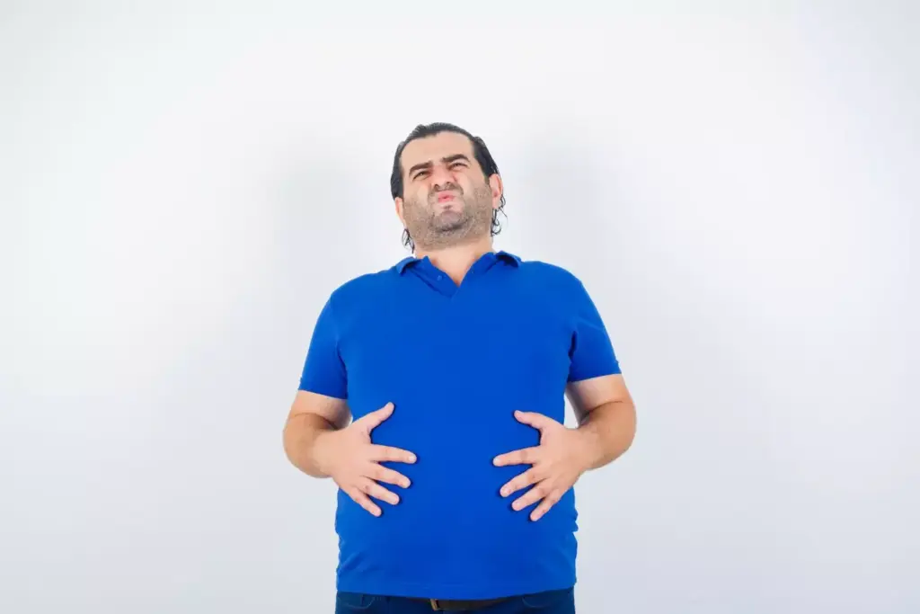 A middle-aged person with a bloated, swollen abdomen lies on their side, face partially obscured. The skin appears red and irritated, with visible stretch marks and a protruding navel. The lighting is soft and diffuse, casting gentle shadows that accentuate the distended midsection. The background is blurred, focusing attention on the distressed, uncomfortable-looking belly. The overall mood is one of concern and unease, reflecting the discomfort experienced by cancer patients during chemotherapy.