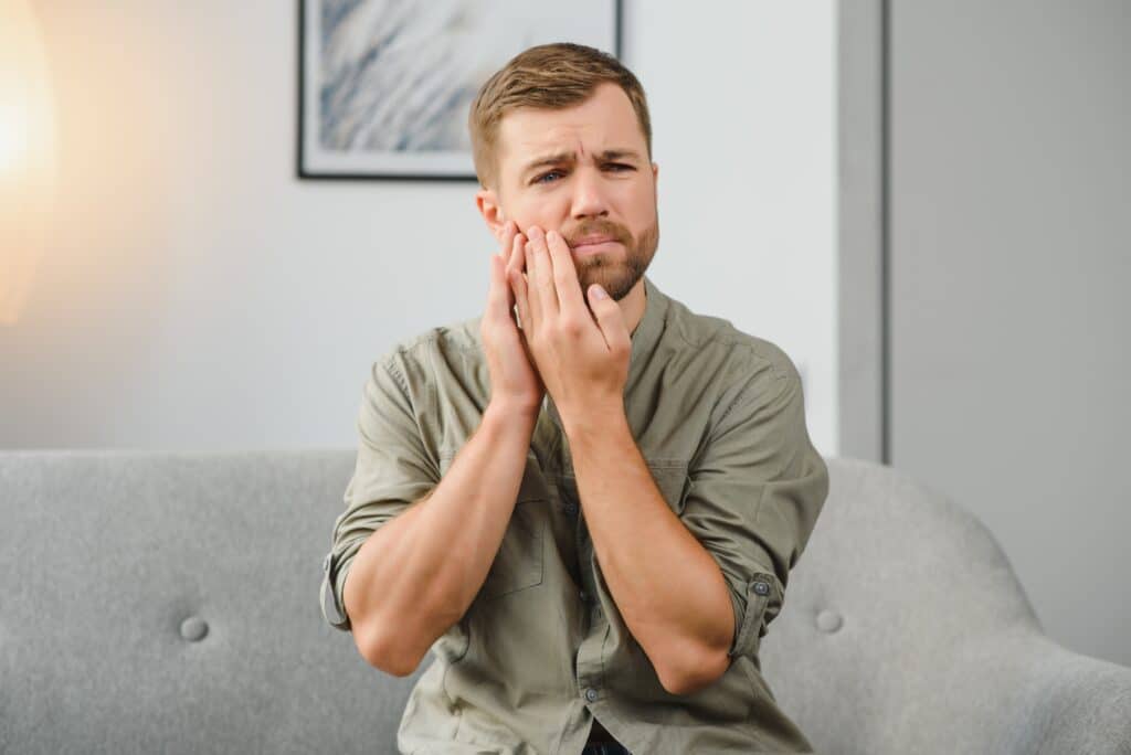 dental health problems concept man suffering from strong toothache upset male patient with inflamed nerve tooth abscess teeth sensitivity sitting sofa touching cheek with grimace pain LIV Hospital